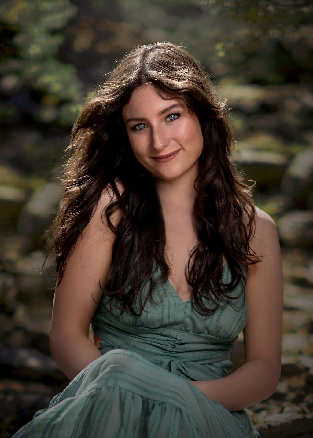 A high school grad sits in a forest in a green dress smiling with hands in her lap