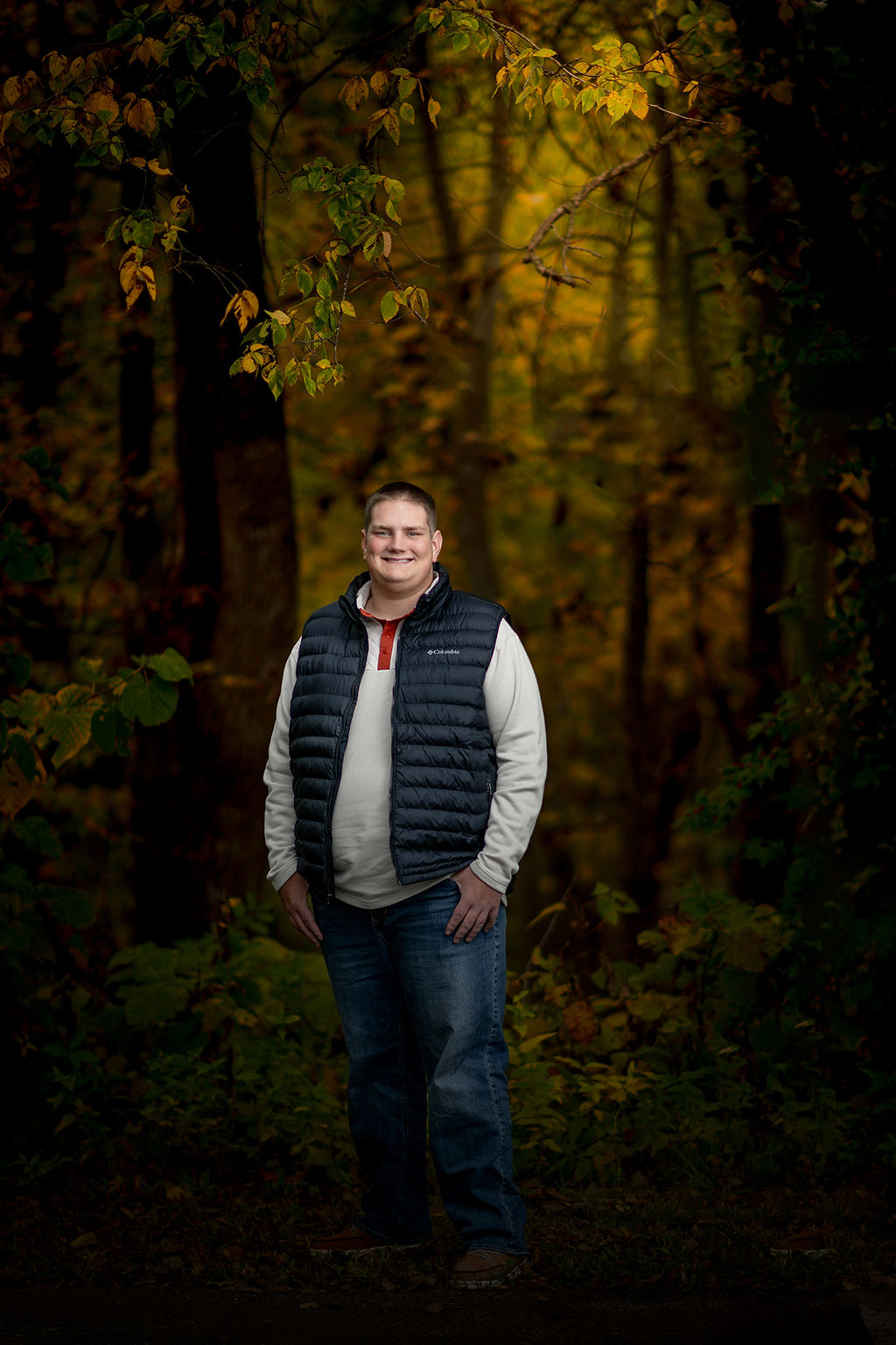 A high school grad stands in a forest in a vest and white shirt with thumbs in his jean pockets after some act prep in des moines