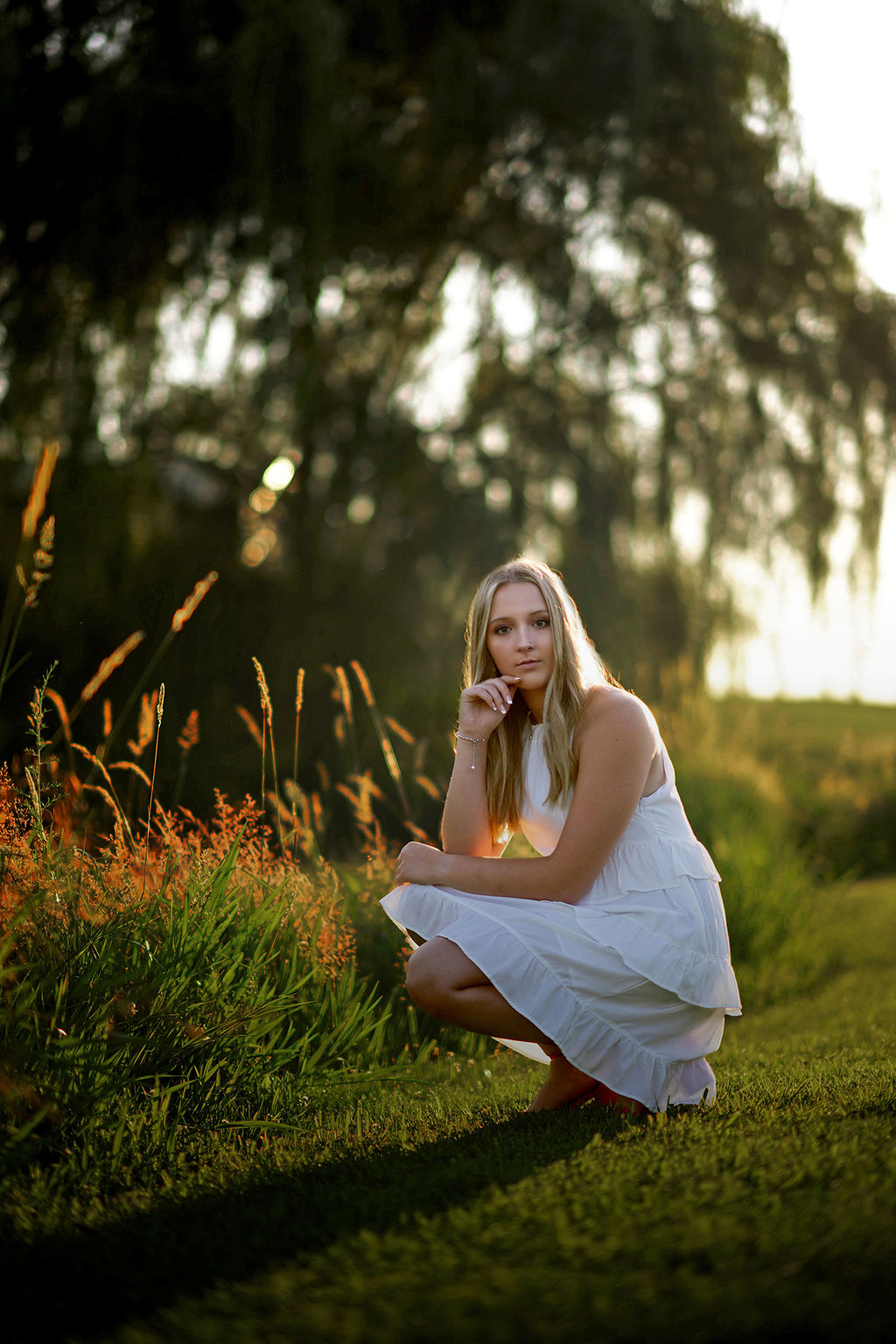 A woman kneels by some flowers in a park at sunset in a white dress