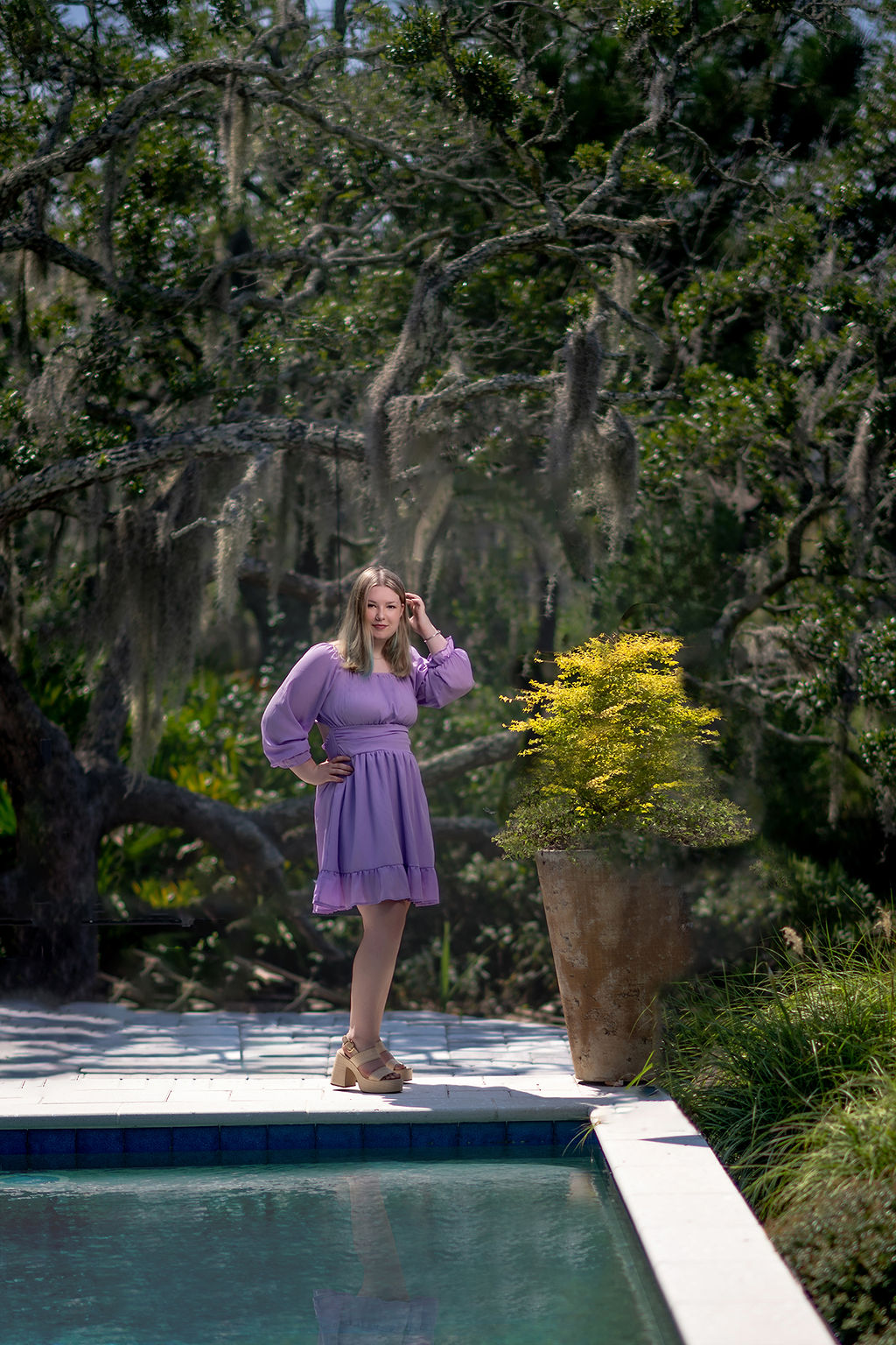 A woman in a purple dress stands by a pool under oak trees with a hand on her hip after visiting boutiques in pella iowa