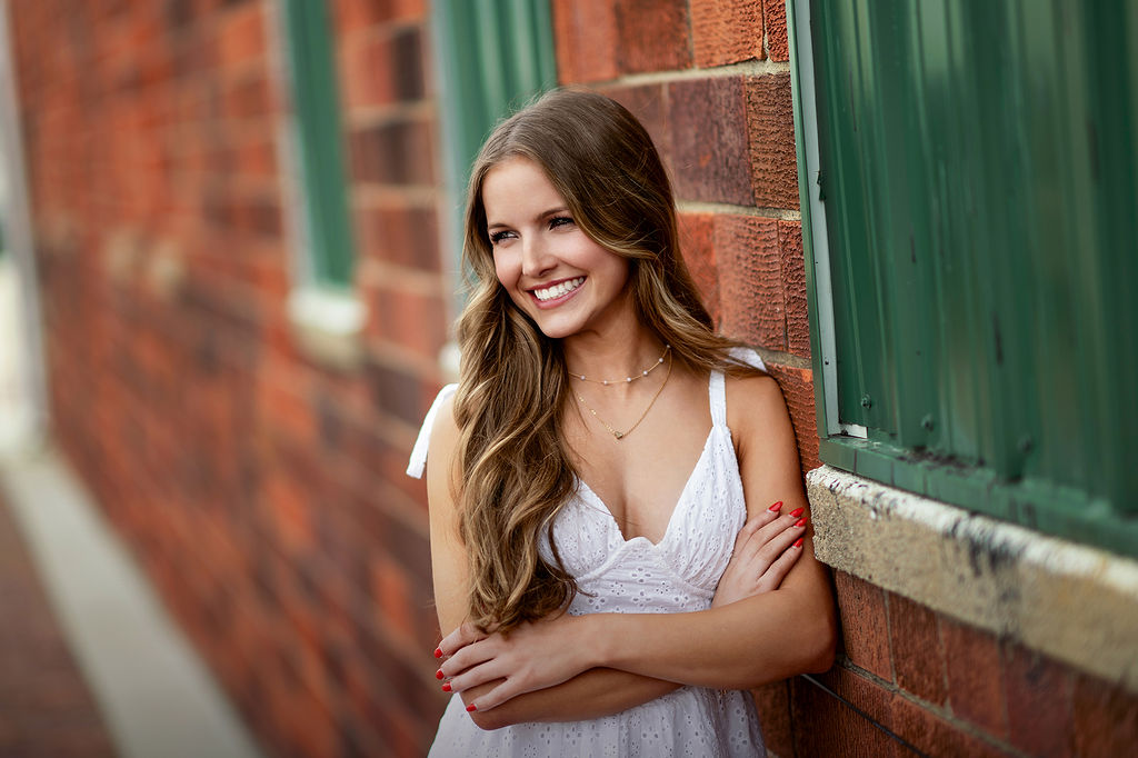 A high school senior leans on a wall while smiling in a white dress with arms crossed after visiting boutiques in pella iowa