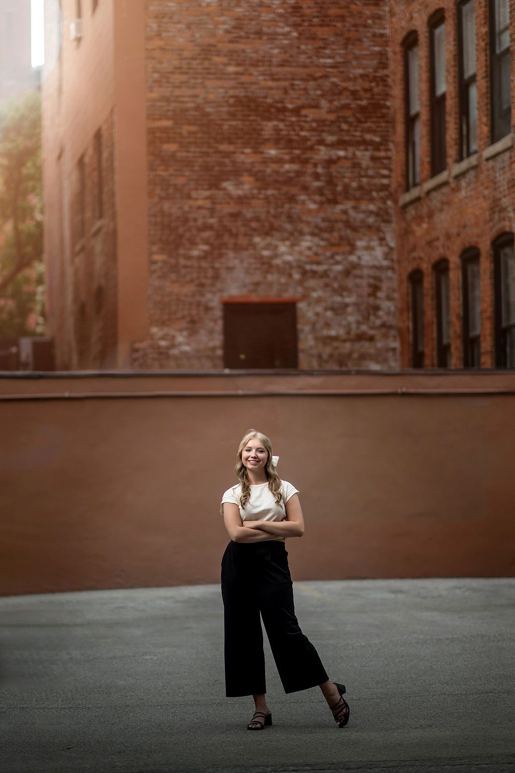 A high school senior in black pants and white shirt stands in a parking lot alley smiling with arms crossed