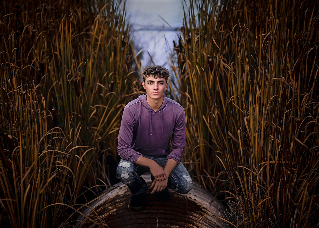 A high school senior squats in a field of tall grasses in a purple hoodie and jeans after visiting central college in pella