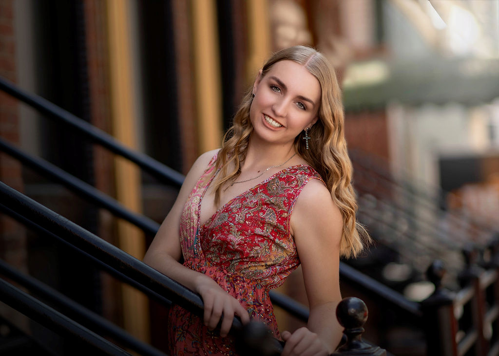 A blonde high school senior in a red print dress walks up stoop steps holding the metal railing after visiting central college in pella