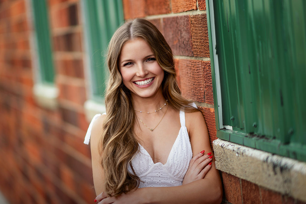 A high school senior in a white dress leans on a brick wall while smiling after exploring colleges near des moines