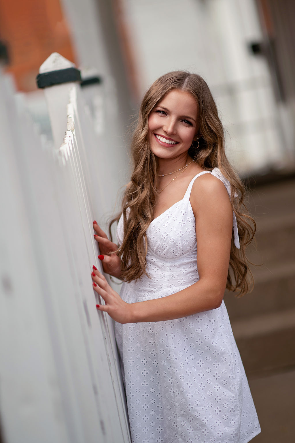 A happy high school senior wearing a white dress leans on a white picket fence smiling