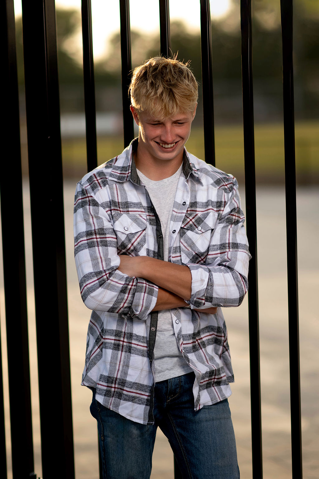 A laughing high school senior in a plaid shirt leans on a black metal fence at sunset