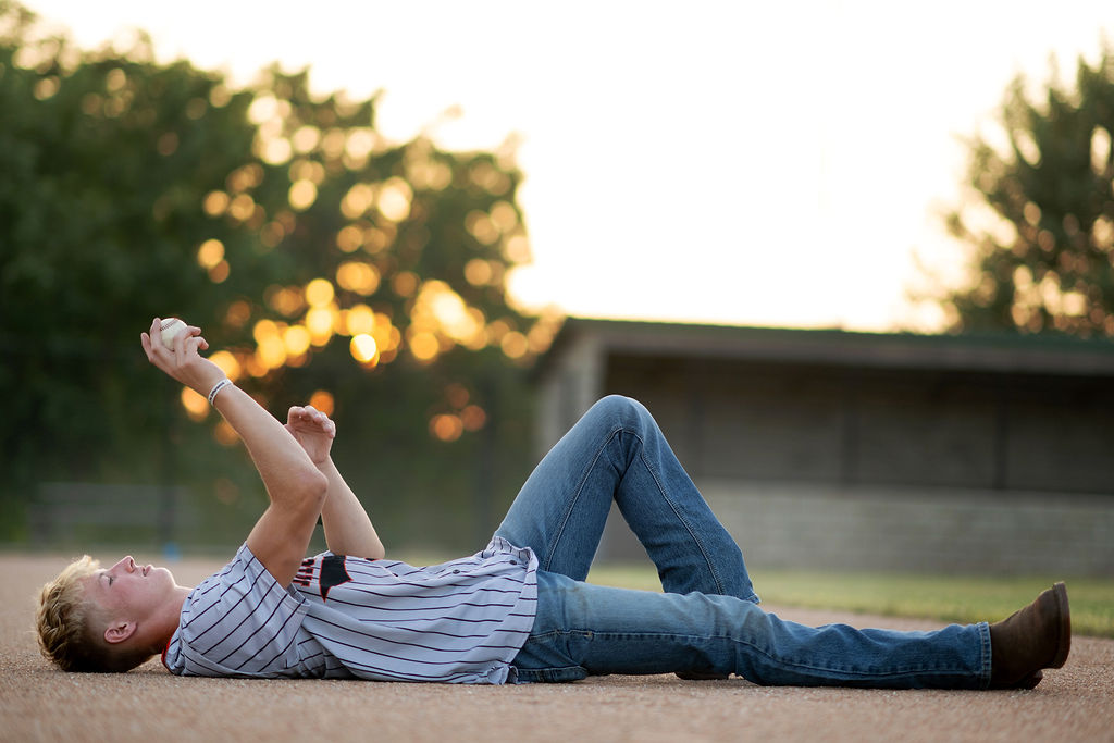 A baseball player lays in the dirt tossing a ball up in the air in jeans and jersey shirt at sunset after using dmacc career advantage