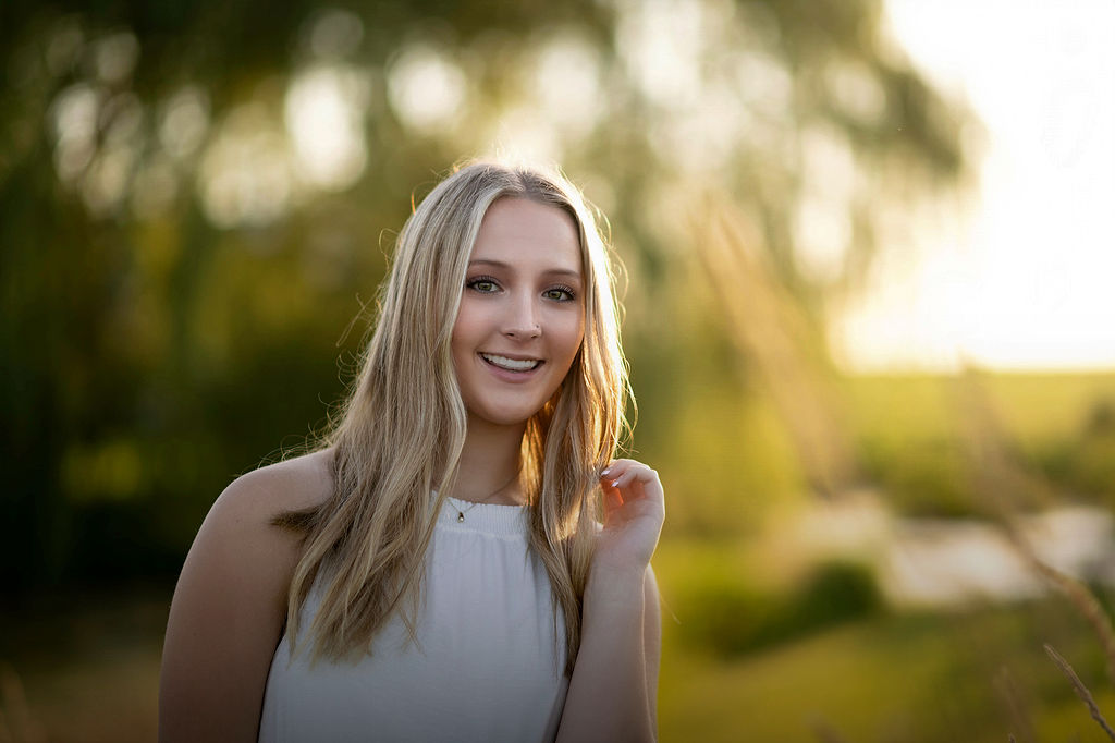 A happy graduate in a white dress stands by a lake at sunset