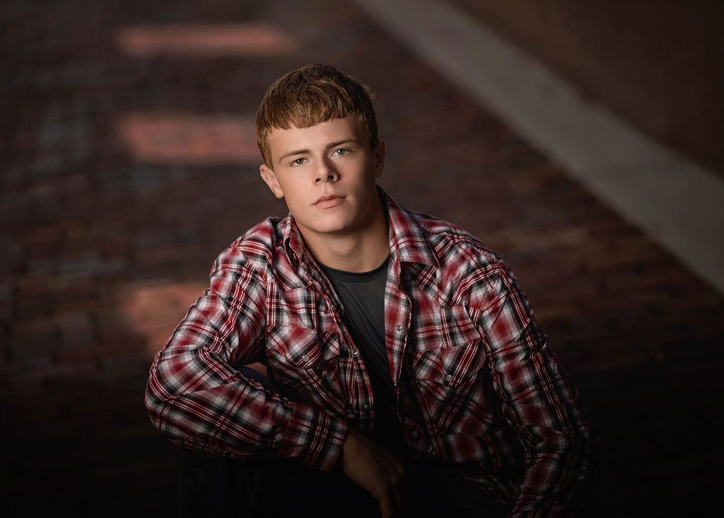 A grad leans on a box in a brick street in a red plaid button down during his drake university graduation photos