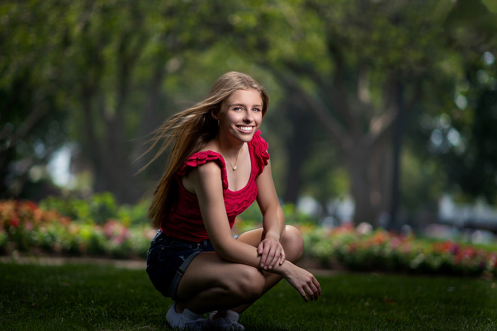 A woman kneels in a garden in a red top and jean shorts with her blonde hair blowing in the wind during drake university graduation photos