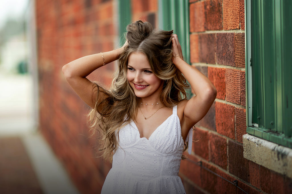 A happy woman in a white dress leans on a brick wall while playing with her hair