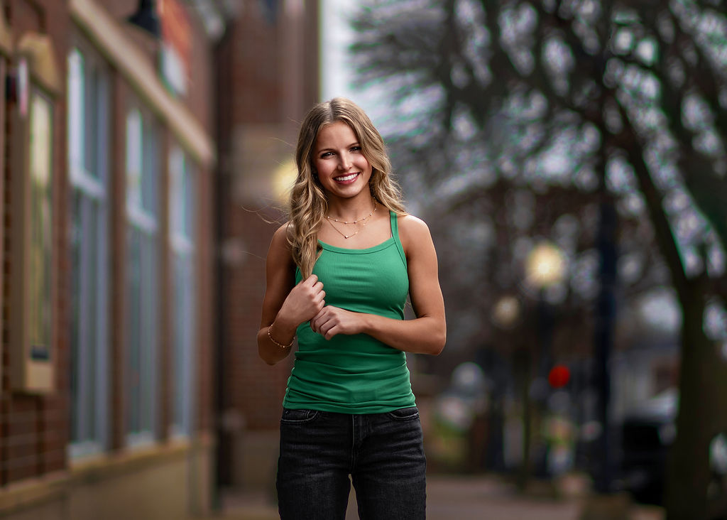 A happy high school senior in a green top plays with her wavy blonde hair while on the sidewalk after visiting hair salons in des moines