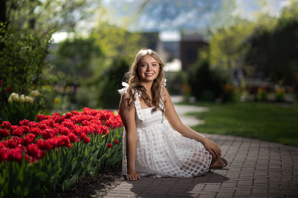 A high school senior in a white dress sits in the sidewalk of a colorful garden smiling after finding homecoming dresses in des moines