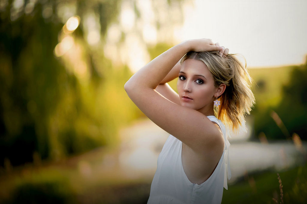 A woman in a white top holds her hair up while standing in a park at sunset for her iowa state graduation photos