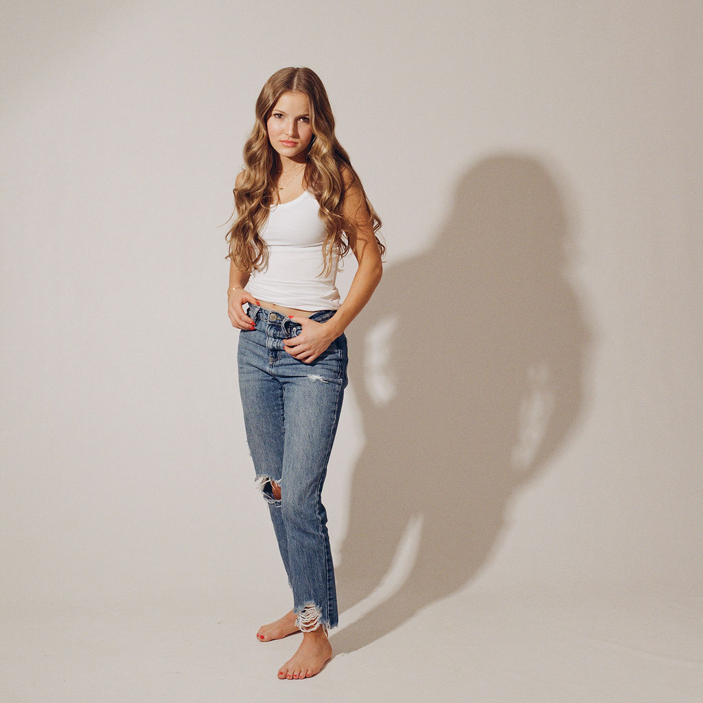 A woman in jeans and white top stands in a white studio with thumbs in her pants