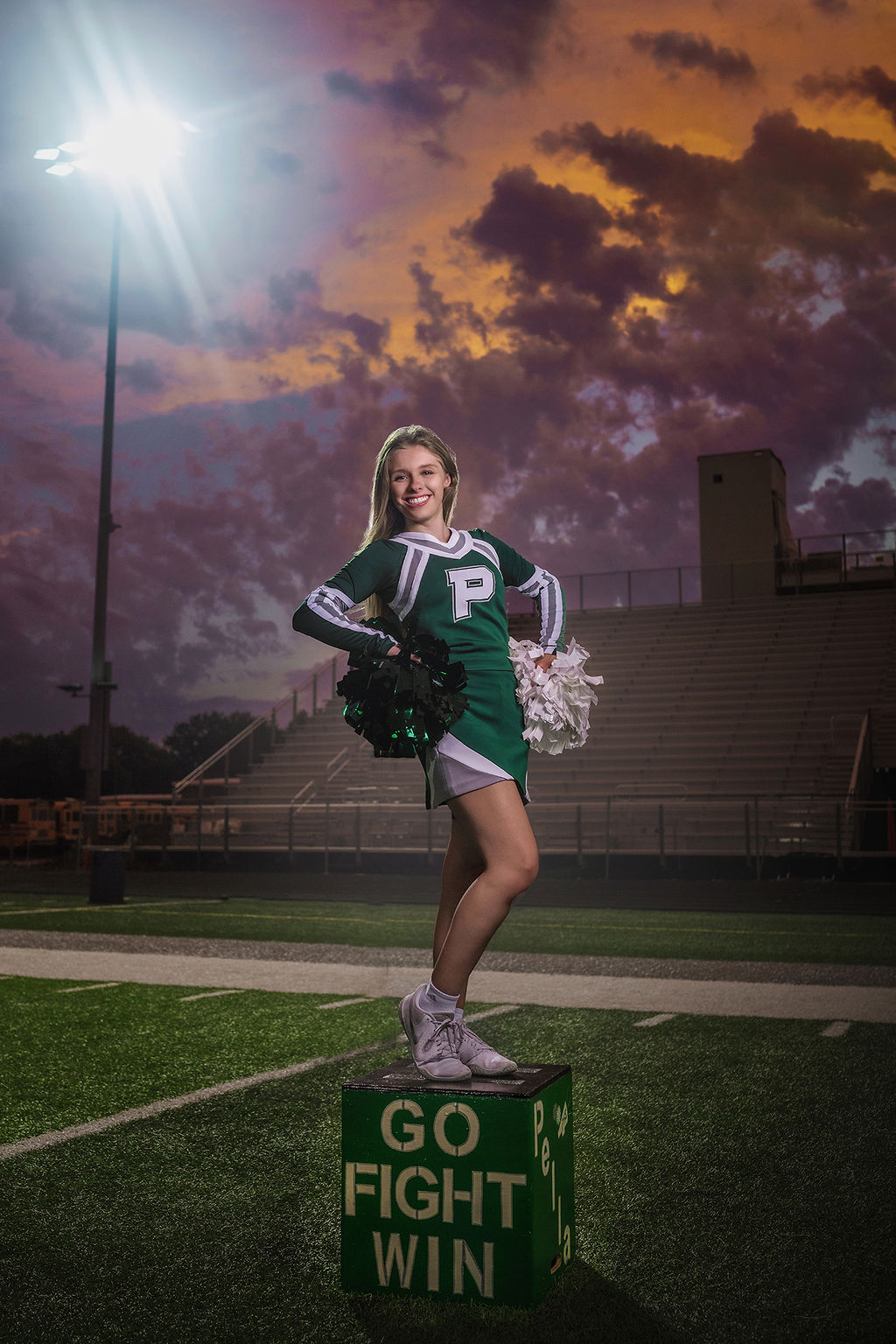 A high school cheerleader stands on a go fight win box on the field under the stadium lights with pom poms on her hips