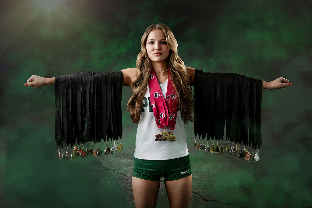 A high school senior stands in a studio showing off her many medals hanging from her extended arms and neck in her pella high school jersey