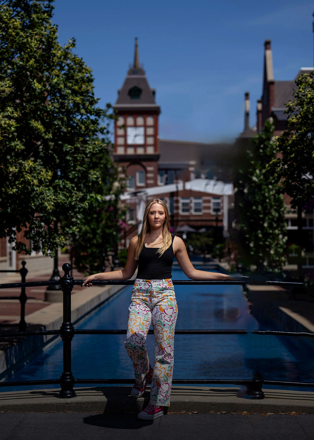 A high school senior in colorful pants and black top leans on a railing by a pool in a couryard