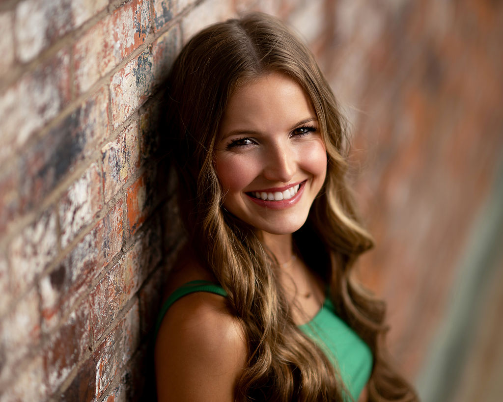 A brunette in a green dress leans on a brick wall smiling after attending private schools in Des Moines