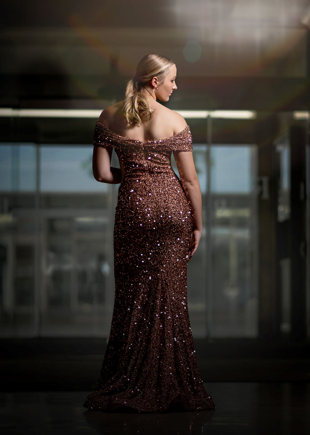 A blonde high school senior stands in a tall glass window in an elegant gown after shopping for prom dresses in Des Moines