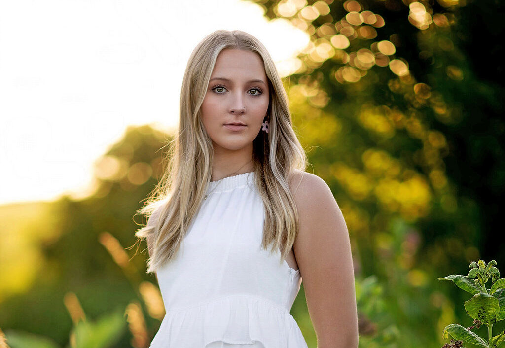 A blonde high school senior stands in a field of tall grasses at sunset in a white dress