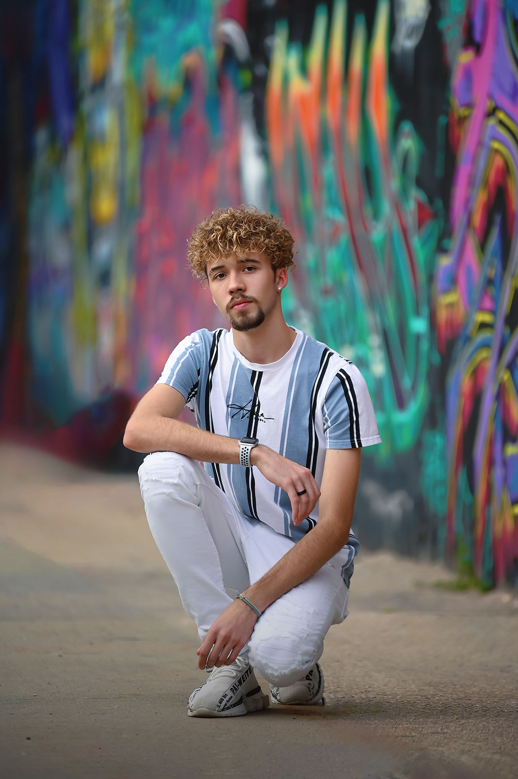 A high school grad kneels on the street in white pants and blue stripe shit by some graffiti