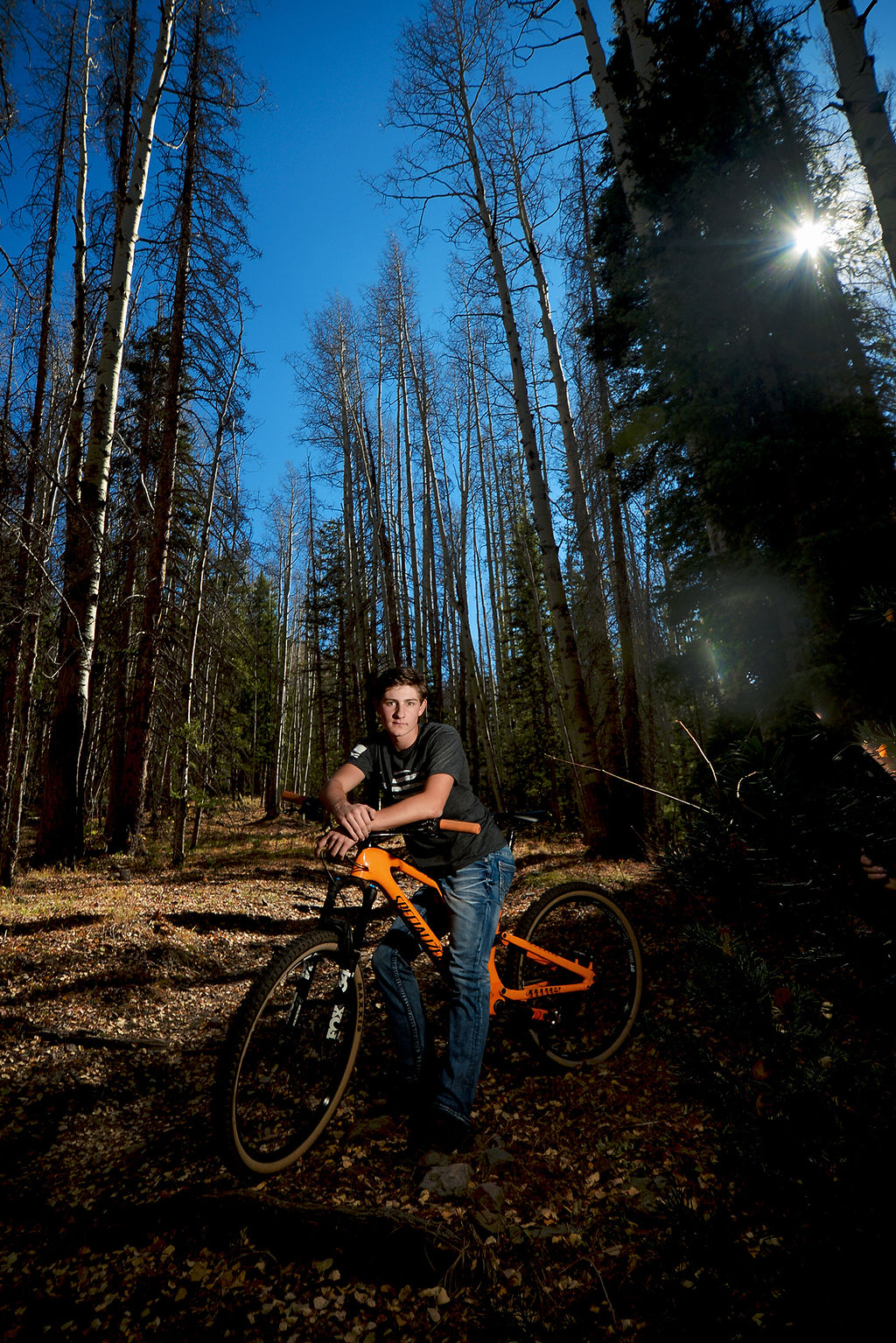 A high school senior sits on his mountain bike in the forest in jeans and black shirt