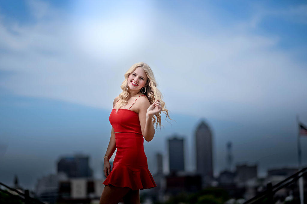 A high school graduate walks in a park overlooking the city in a red dress for her senior photos in des moines