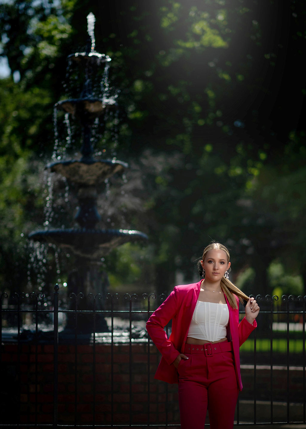 A high school senior in a white top and hot pink jacket and pants leans on an iron fence of a park fountain playing with her hair after visiting tanning salons in des moines