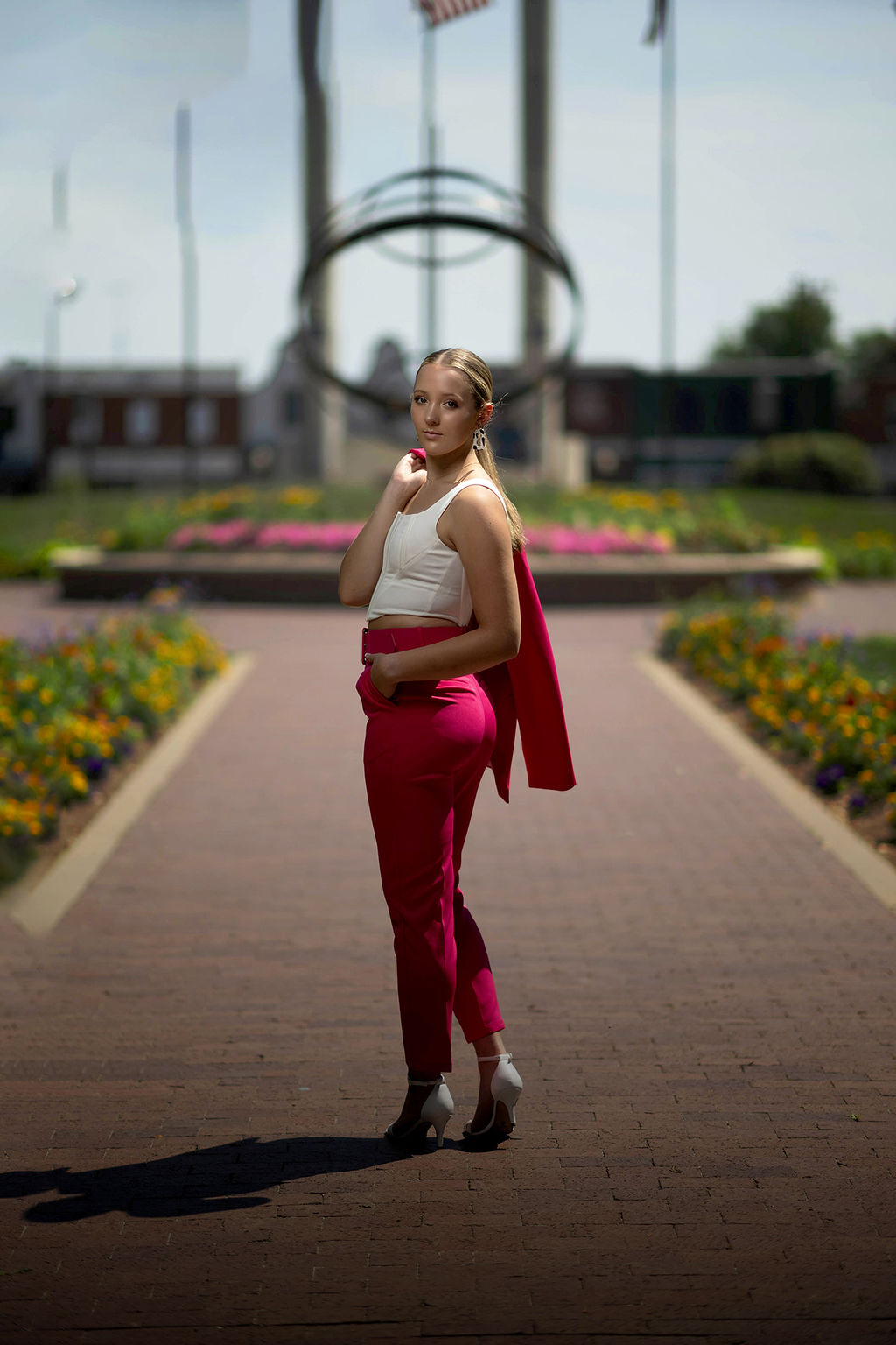 A high school senior in hot pink pants walks on a garden sidewalk while turning to look over her shoulder with a hand in her pocket after visiting tanning salons in des moines