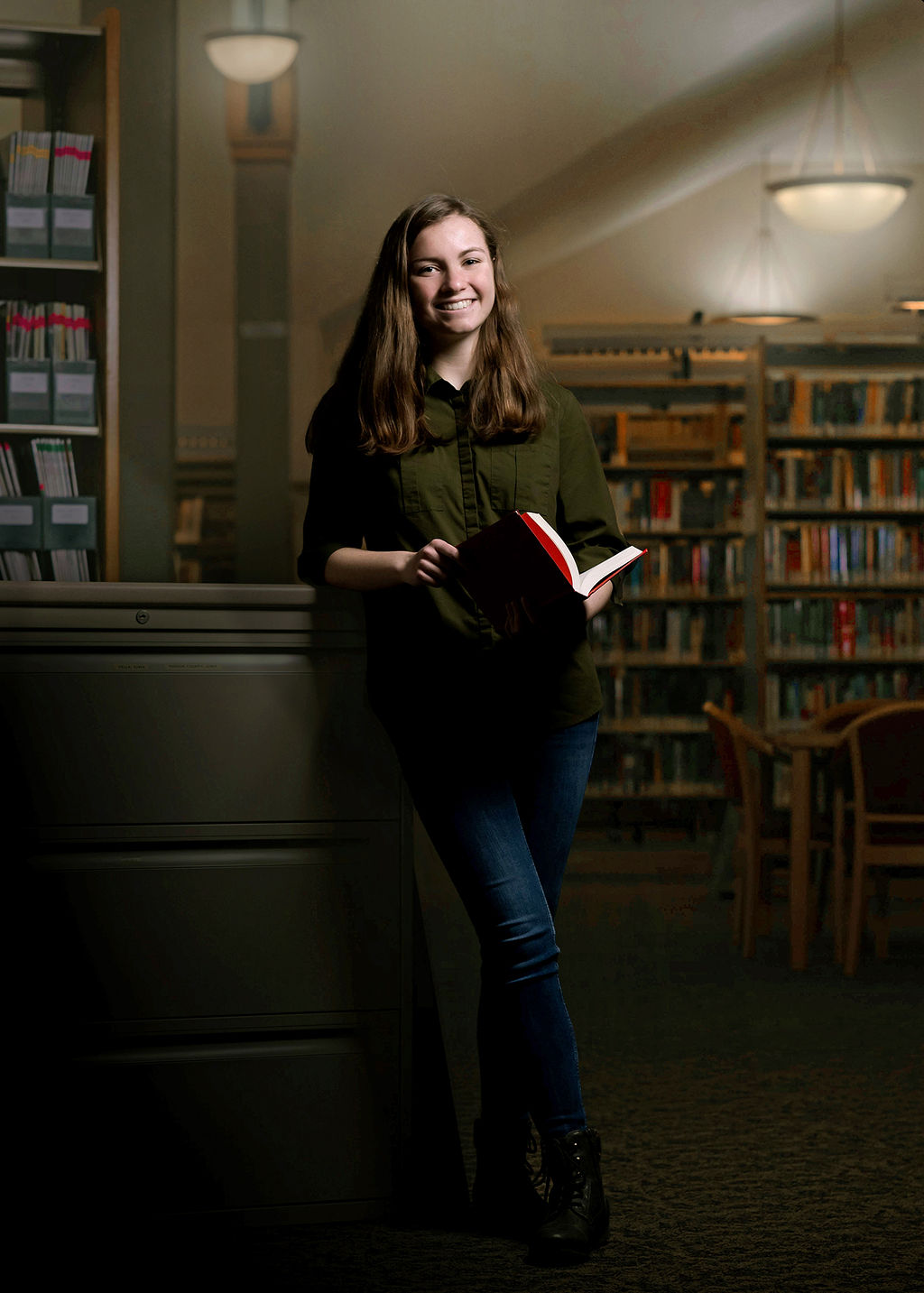 A high school senior stands in a library with a book in her hands in jeans and green shirt