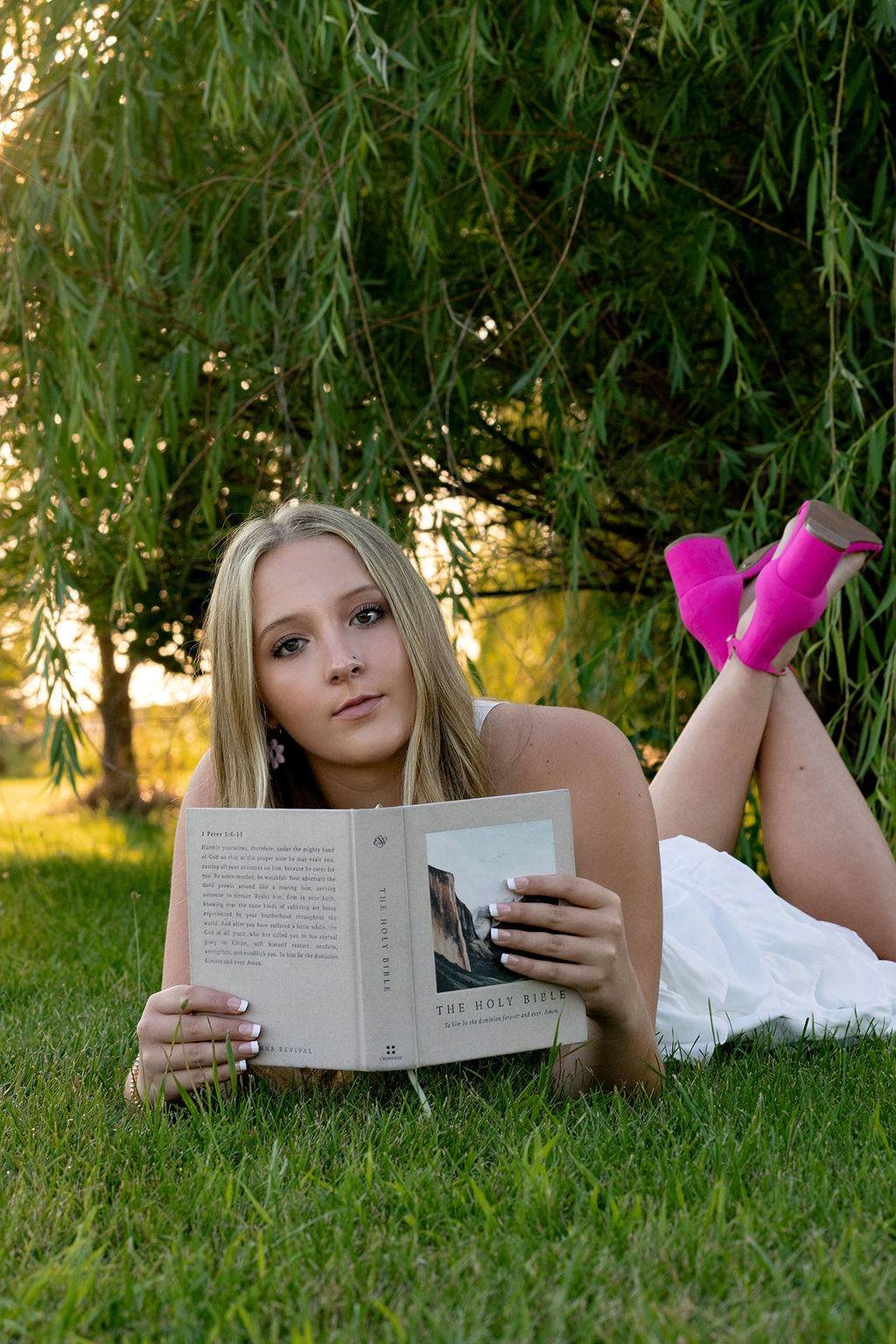 A high school senior lays in the grass under a willow tree at sunset while reading the bible in a white dress and pink shoes after finding tutors in des moines