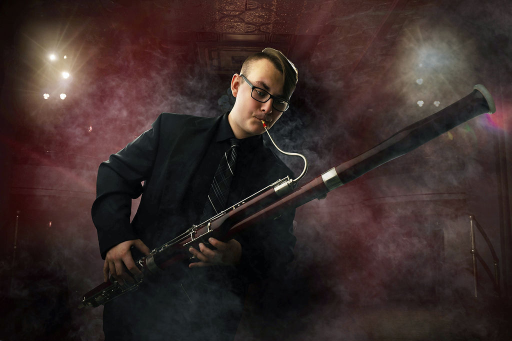 A bassoon player stands on a stage filled with smoke playing in a black suit and tie