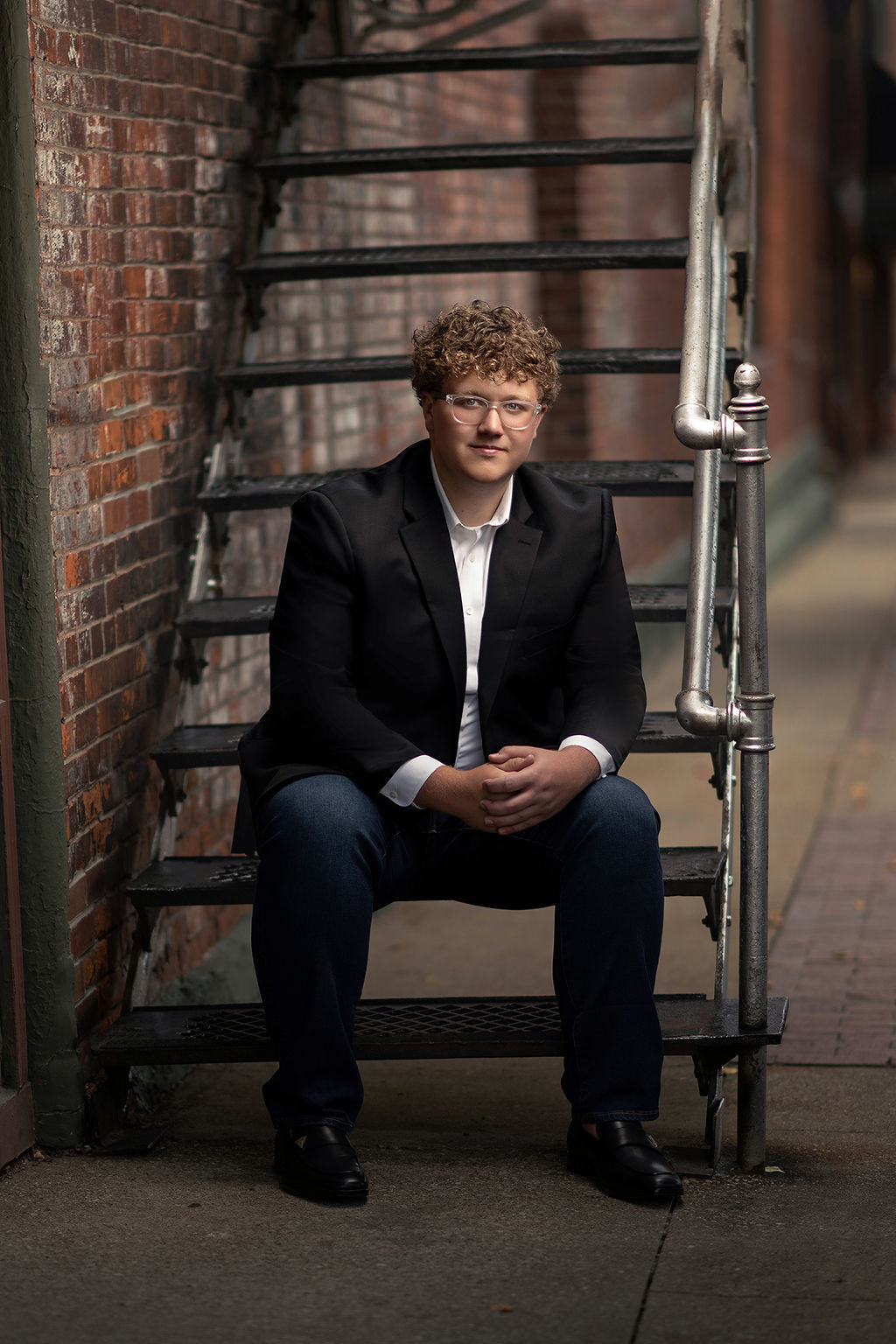 A high school senior sits on a set of metal stairs in an alley in a black jacket and jeans after finding a tux rental in Des Moines