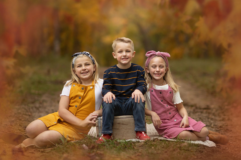 A boy sits on an apple box in a park trail smiling with his two older sisters on either side