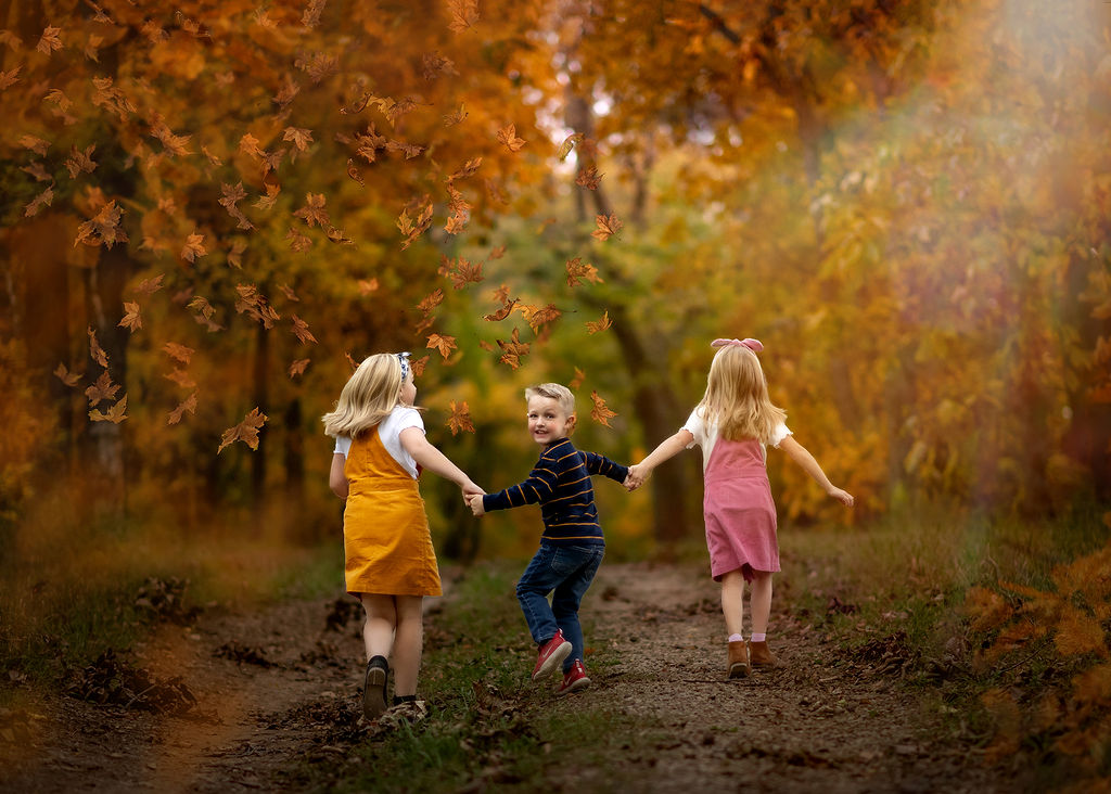 A toddler boy looks over his shoulder while running in a park trail in fall holding hands with his older sisters after visiting Janes Family Shop