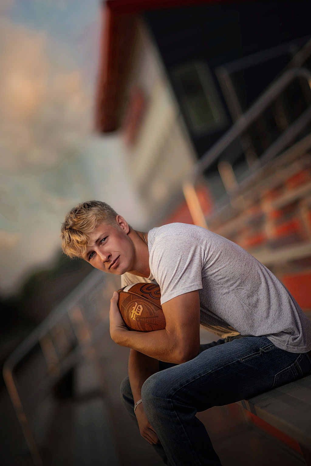 A high school senior sits on bleachers holding a football in one arm in a grey shirt and jeans at sunset at Oskaloosa High School