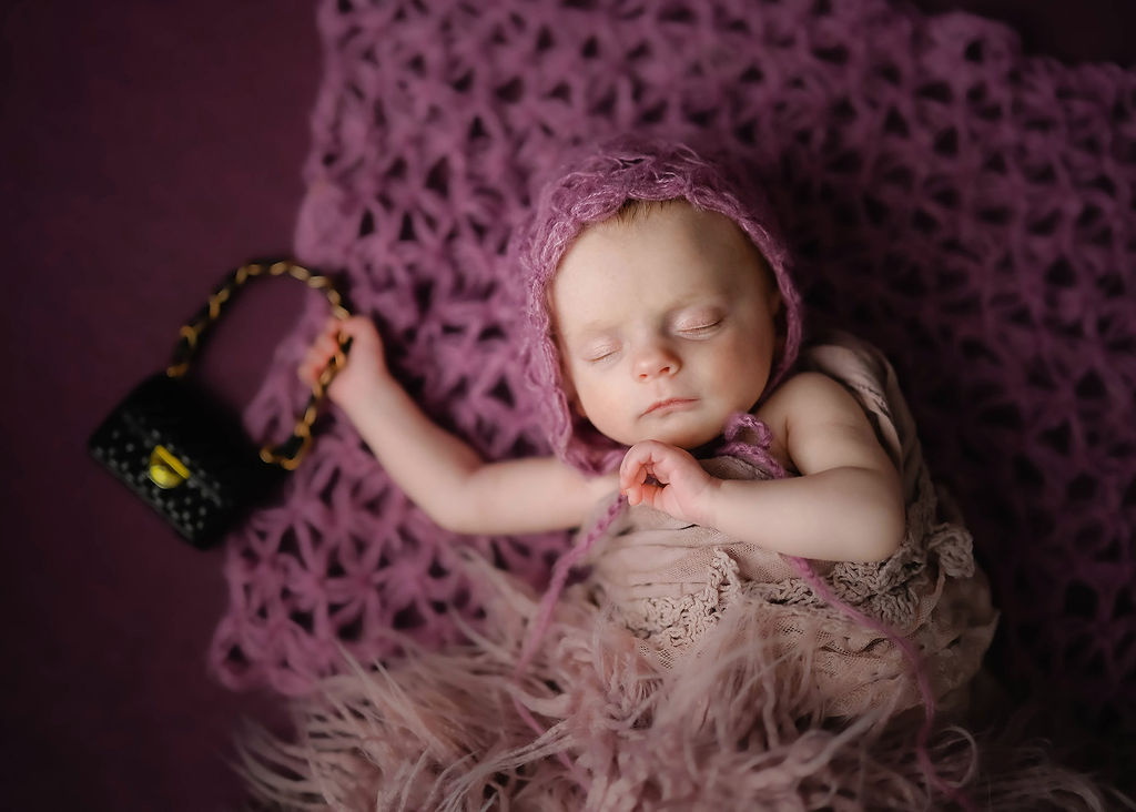 A newborn baby sleeps holding a tiny purse in a brown blanket and knit bonnet