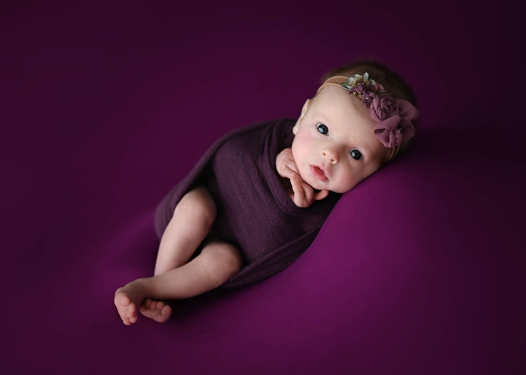 A newborn baby lays on a purple bed with eyes wide open in a matching swaddle after visiting a baby boutique in des moines