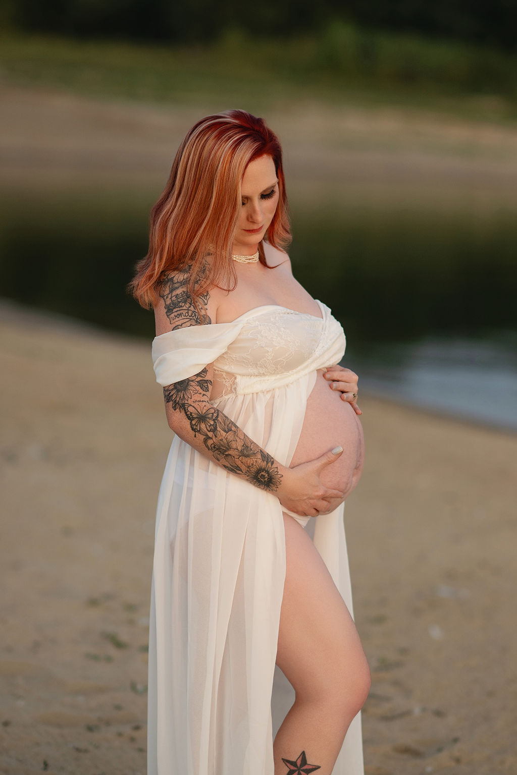 A mother to be holds her exposed bump on a beach in a white maternity gown at sunset