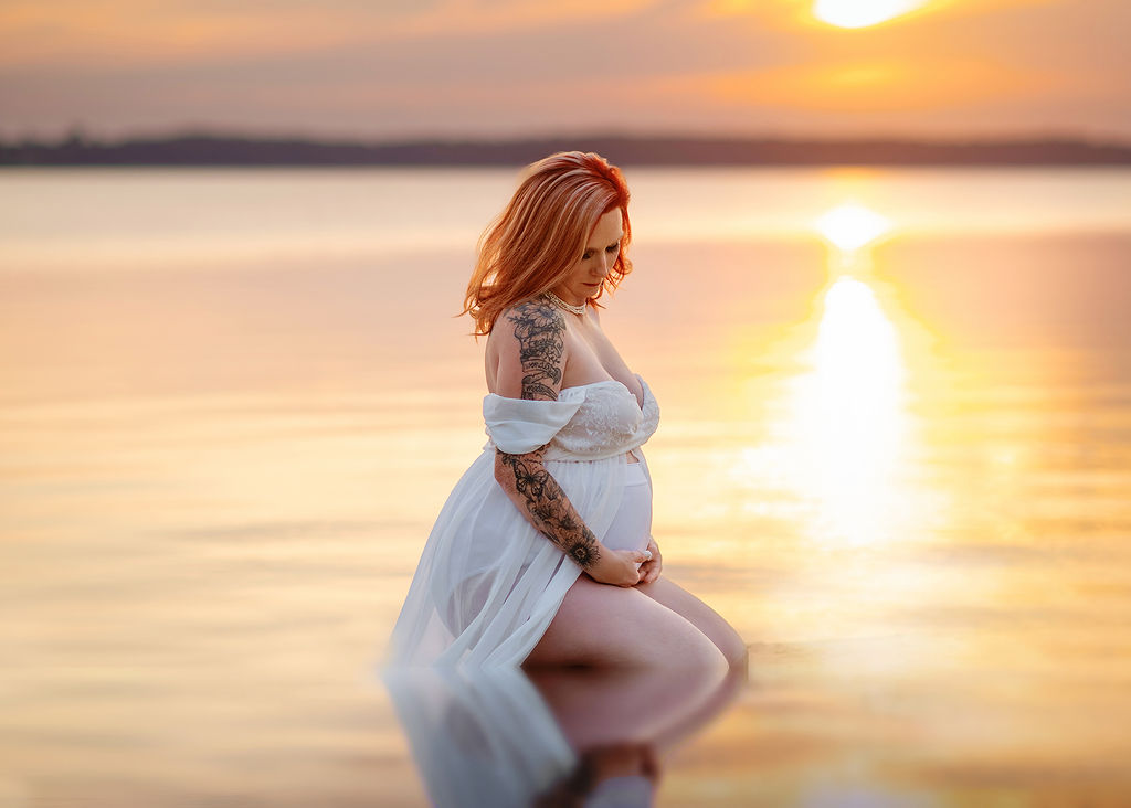 A mother to be kneels in the water at sunset in a white maternity gown looking down at her bump after visiting baby shower venues in Des Moines