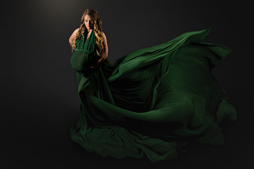 A pregnant woman stands in a studio as her green maternity gown flows behind her