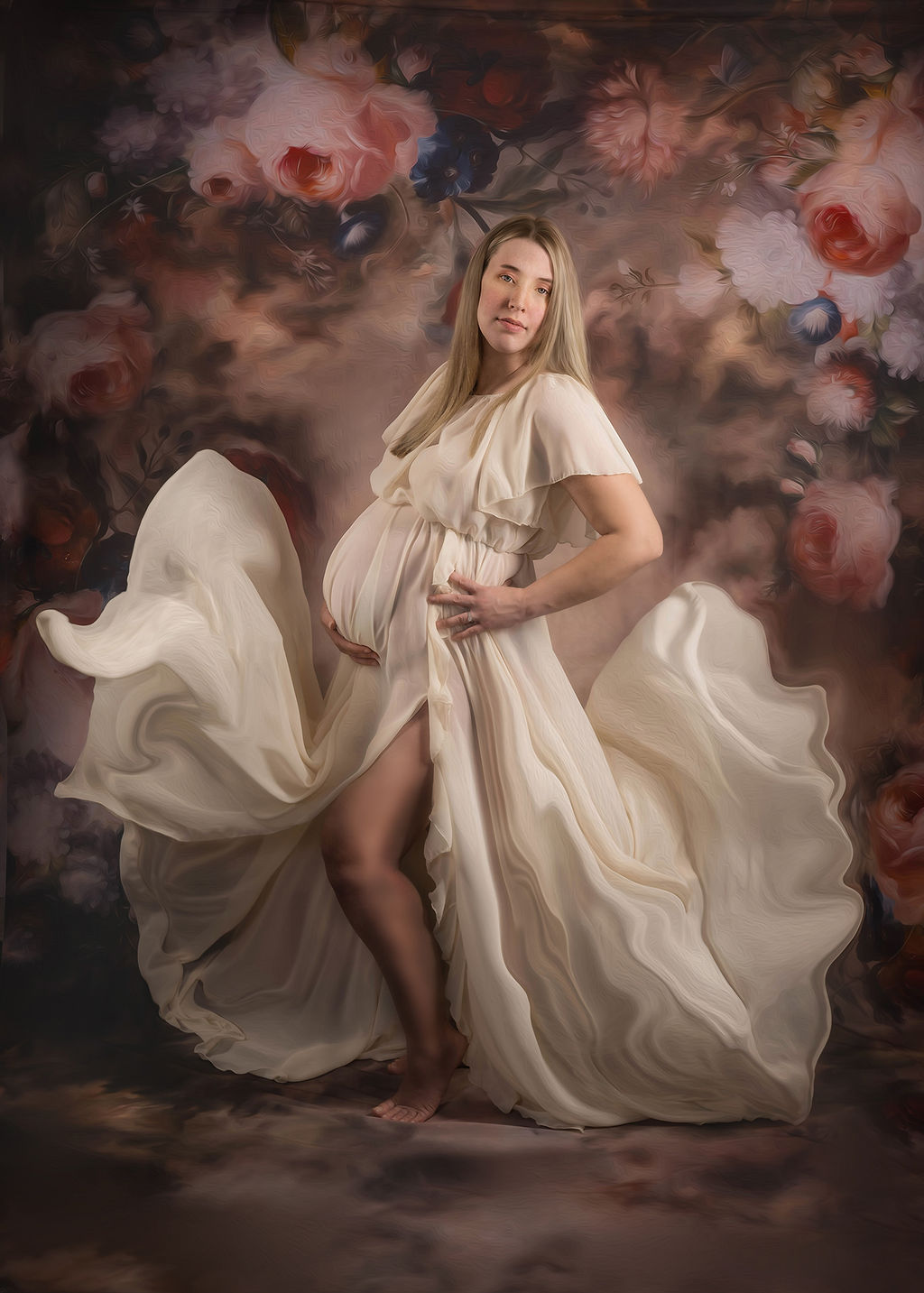 A mom to be stands in a studio in a white maternity gown as it flows around her with a floral backdrop after visiting a birthing center in des moines