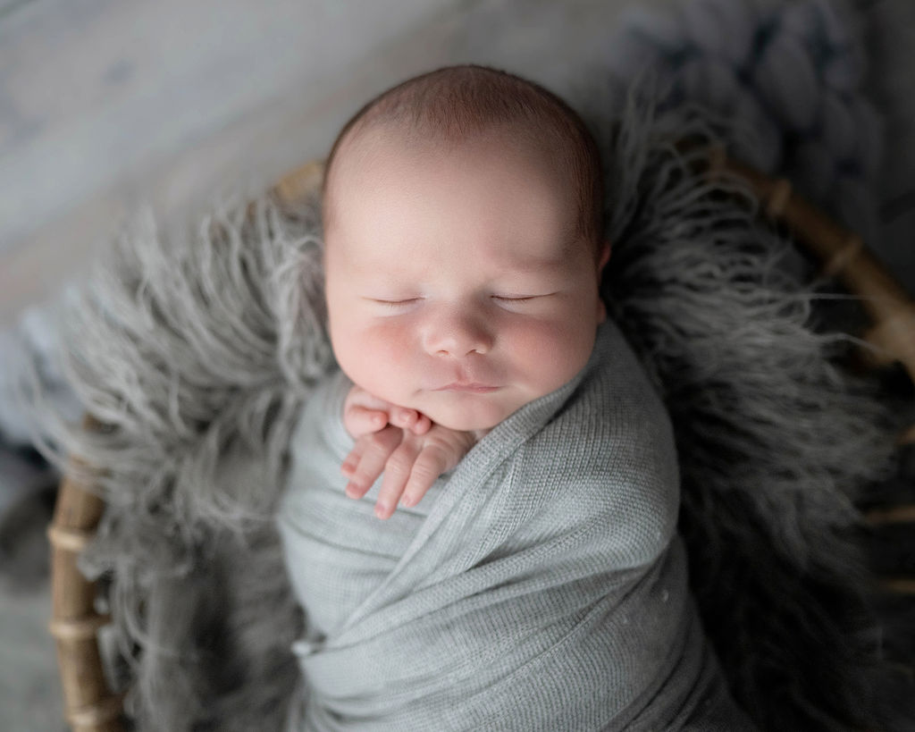 a newborn baby sleeps in a grey swaddle in a wicker blanket in a studio after visiting Daycares in Des Moines