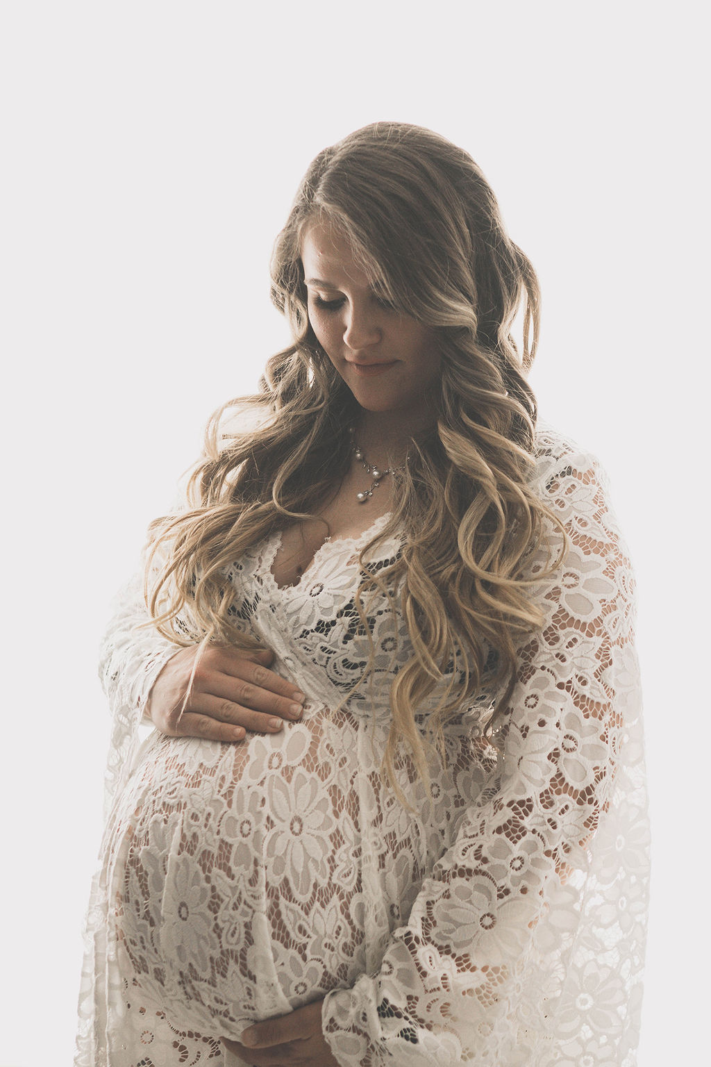 A mom to be in a lace maternity gown stands in a studio smiling down at her bump after meeting des moines midwives