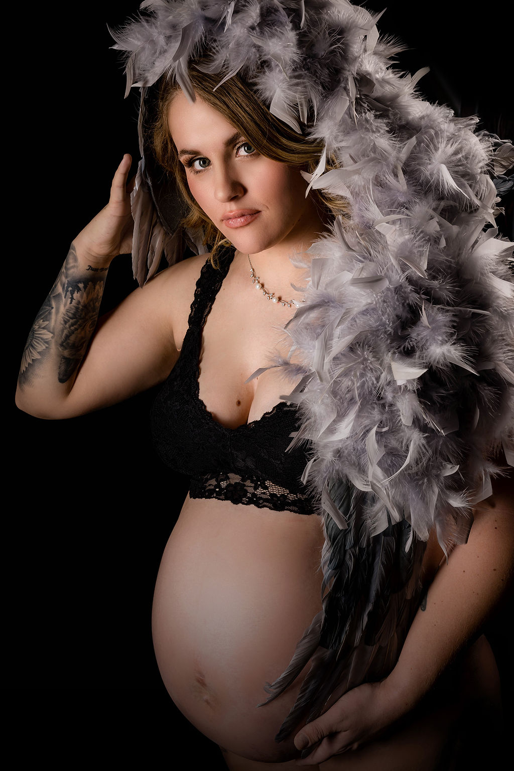 A pregnant woman stands in a studio covered in feathers in a black lace bra