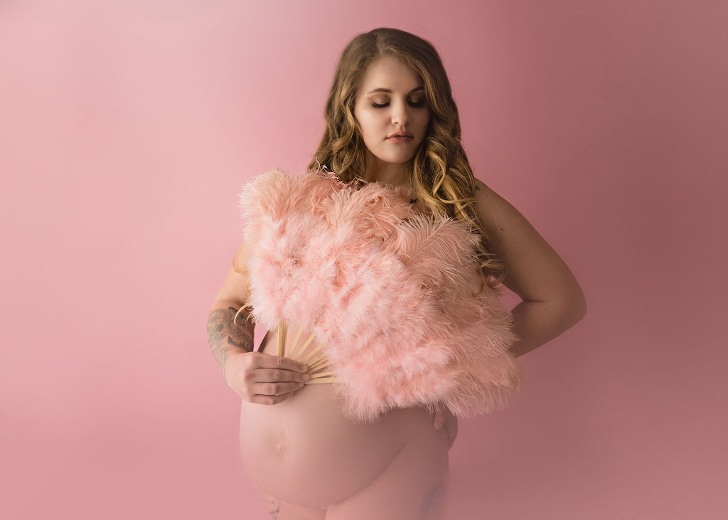 A mom to be covers with a pink feather fan while gazing down her shoulder in a pink studio after visiting family birthing center pella