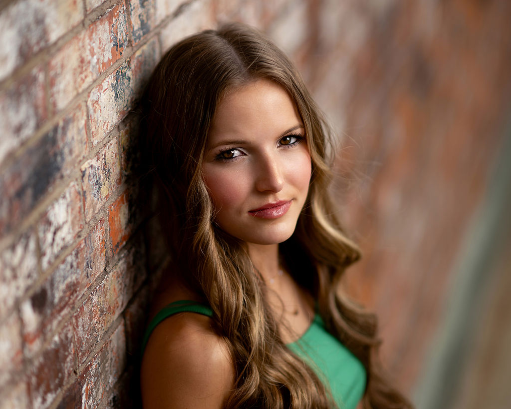 A high school senior leans on a brick wall in a green top