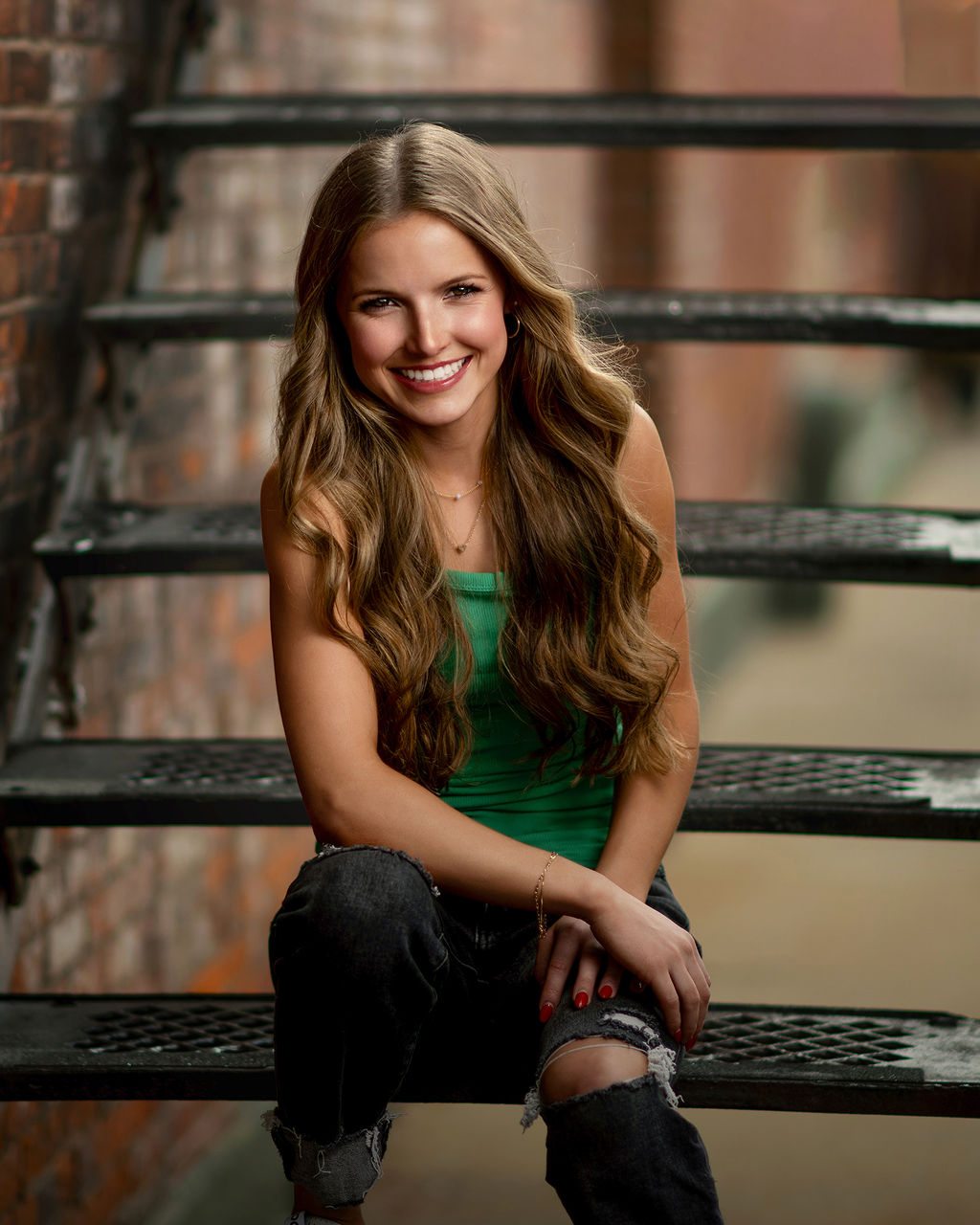 A laughing grand view university graduation girl in a green top and ripped jeans sits on metal alley stairs